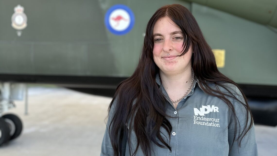 Maddie Ward has long dark hair and is smiling. She is wearing an Endeavour Foundation collared shirt and standing in front of an RAAF aircraft.