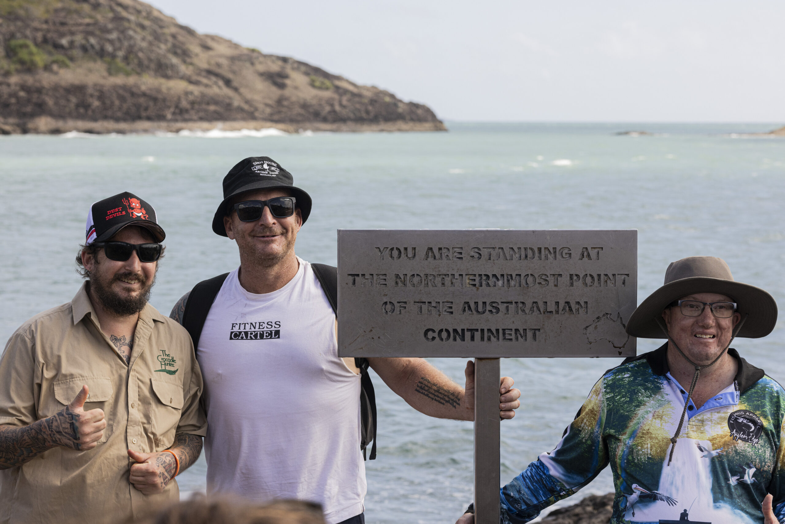 Mark Swift, Endeavour Foundation, Employment Coach is standing between two people we support in front of a sign saying, You are standing at the northernmost point of the Australian Continent. All three men are smiling, two people we support have their thumbs up.