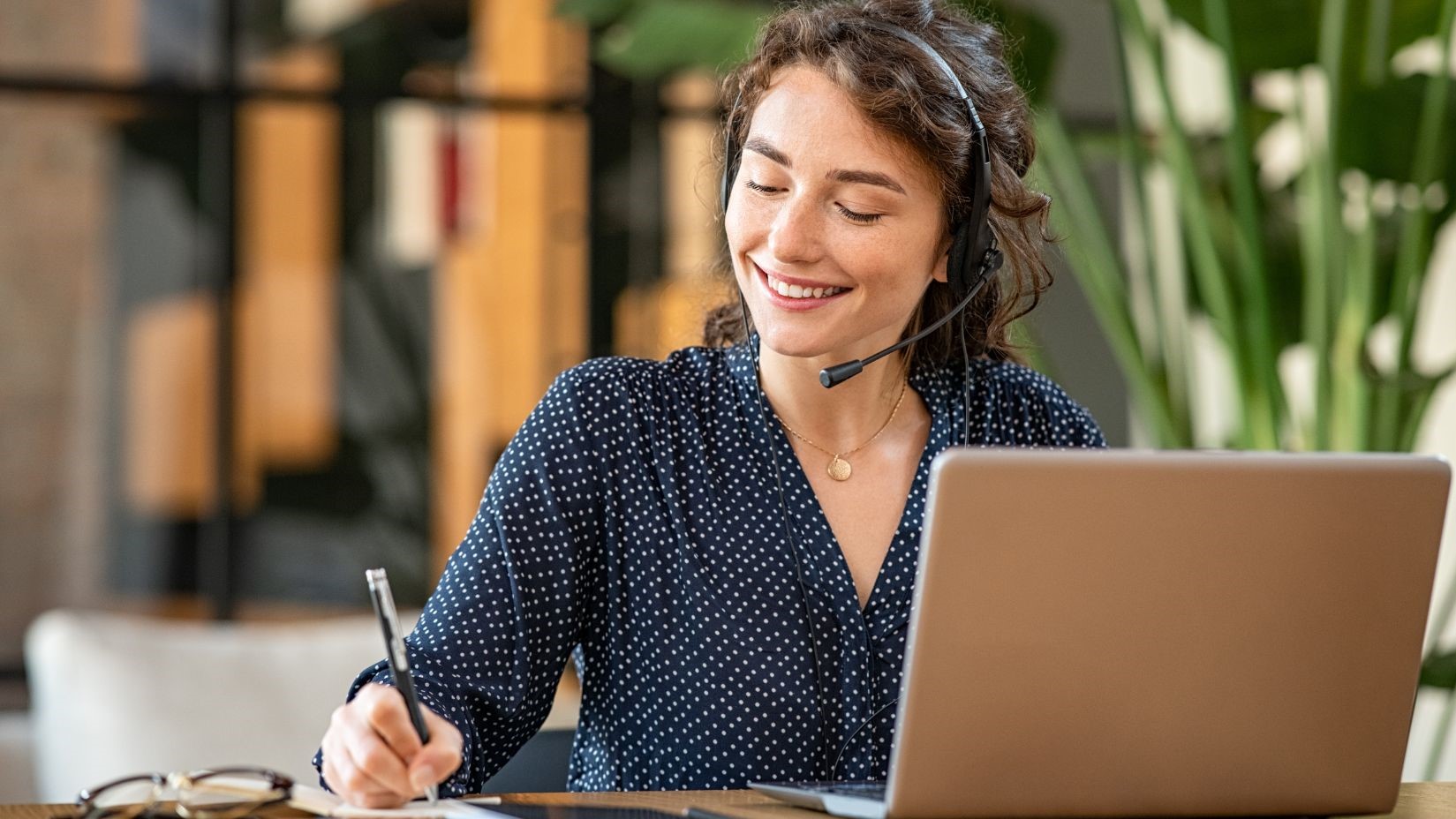 Women who is smiling and talking to a person on a computer and taking notes.